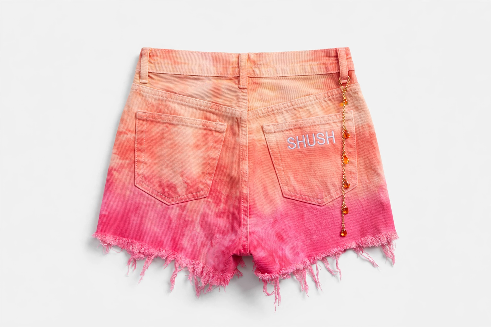 Women's tie-dye denim cutoff shorts in a peach-to-hot-pink sunset ombre wash, SHUSH embroidered in white on the back pocket, gold chain with orange gem charms at the belt loop